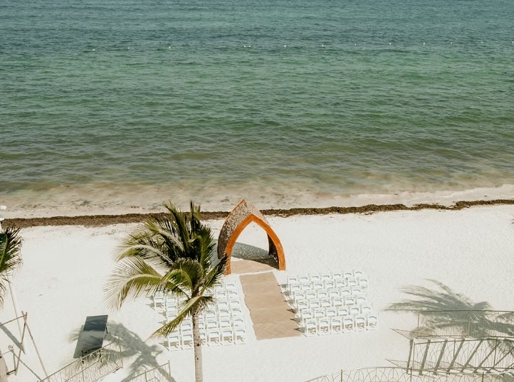 screenshot 20260713 164230 be all inclusive Beach ceremony setup with an orange arch, white chairs in rows, and an aisle leading to the arch overlooking the ocean