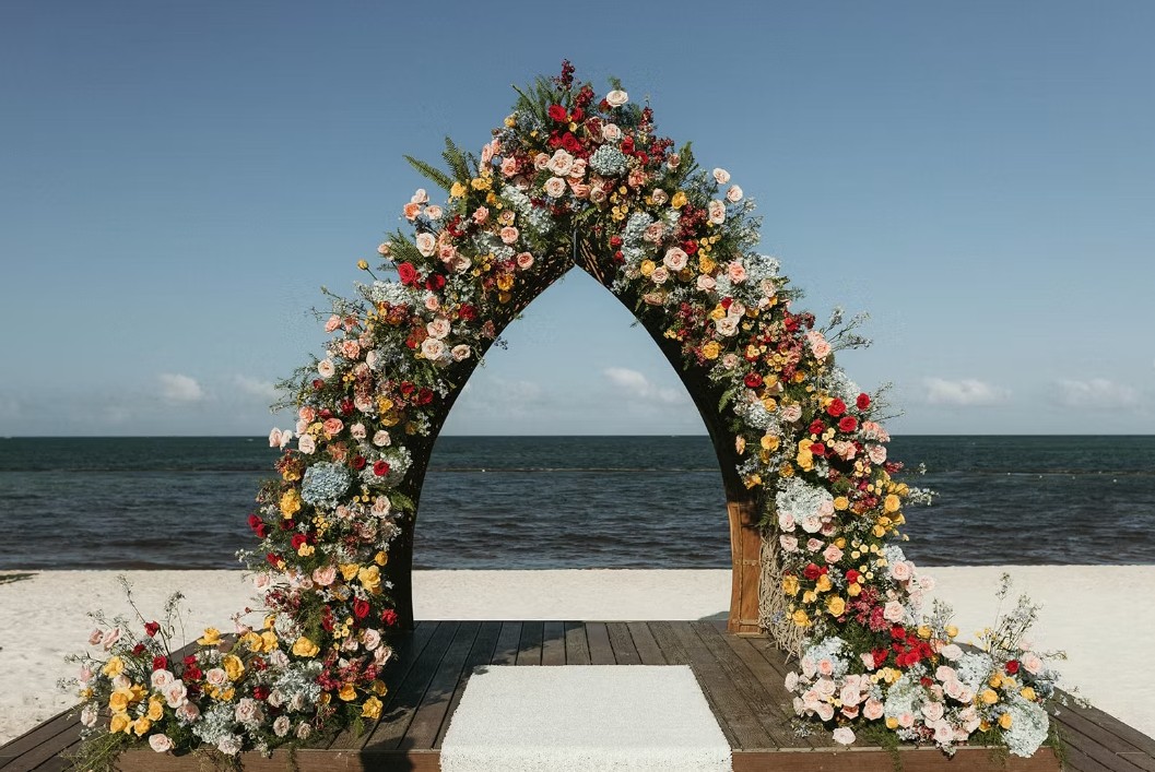 screenshot 20260713 164243 be all inclusive Floral arch on a beach ceremony platform with colorful roses and greenery against the sea backdrop.