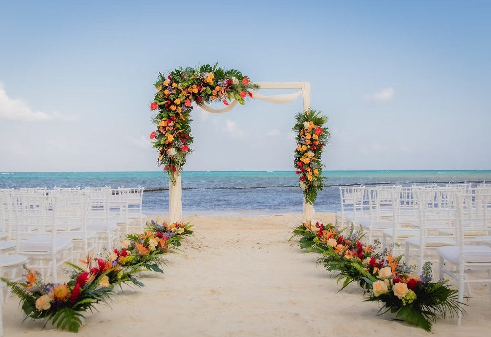 screenshot 20260713 164403 be all inclusive Beach wedding arch decorated with colorful tropical flowers and greenery, set on sand with white chairs facing the sea.