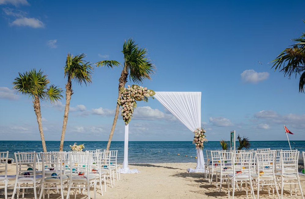 screenshot 20260713 164416 be all inclusive Beach wedding on a sandy shore with a white fabric arch and floral decorations facing the ocean, rows of white chairs await guests.