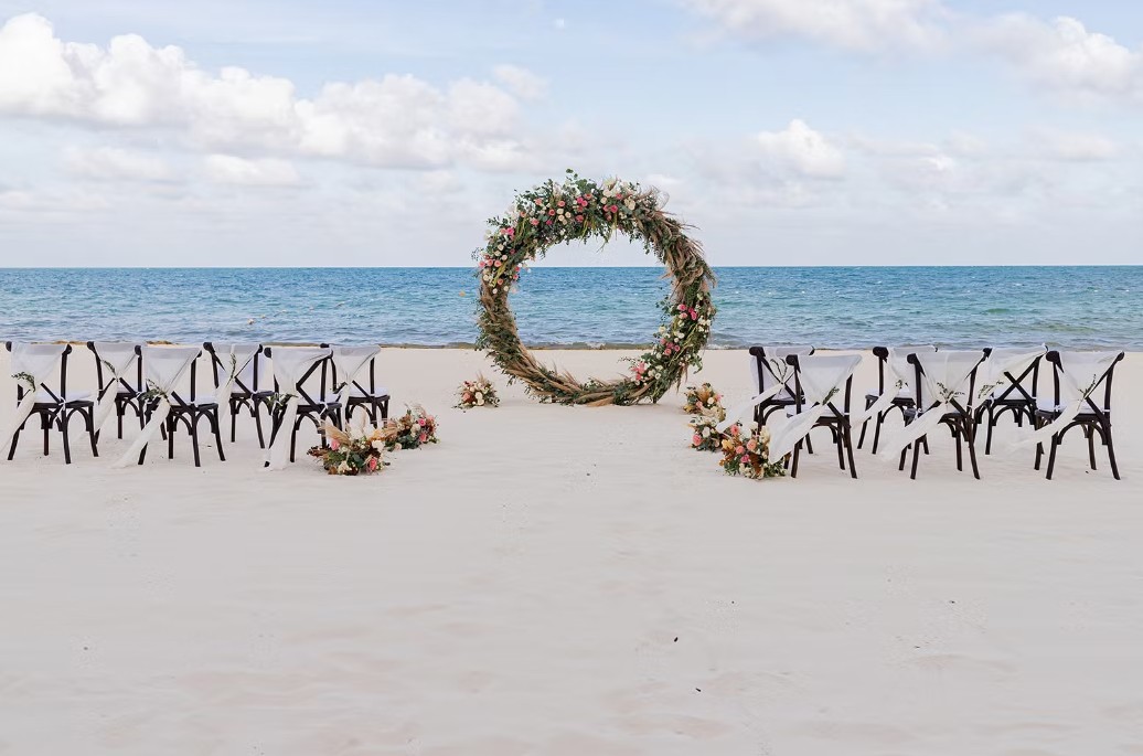 screenshot 20260713 164428 be all inclusive Circular floral wedding arch on a sandy beach with rows of chairs and flower arrangements for an outdoor ceremony by the ocean