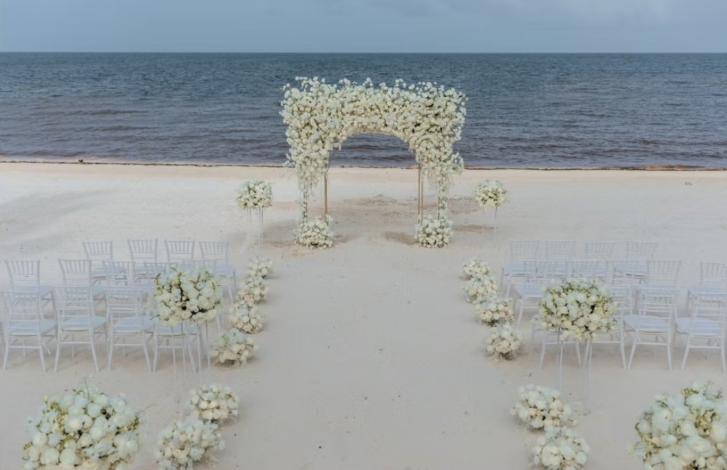 screenshot 20260713 164440 be all inclusive White floral wedding arch on a sandy beach with rows of white chairs facing the arch by the ocean