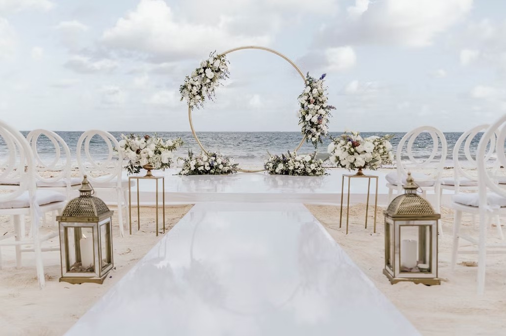 screenshot 20260713 164458 be all inclusive Beach wedding setup with a circular flower arch at the end of a white aisle, lanterns, and white chairs by the sea.