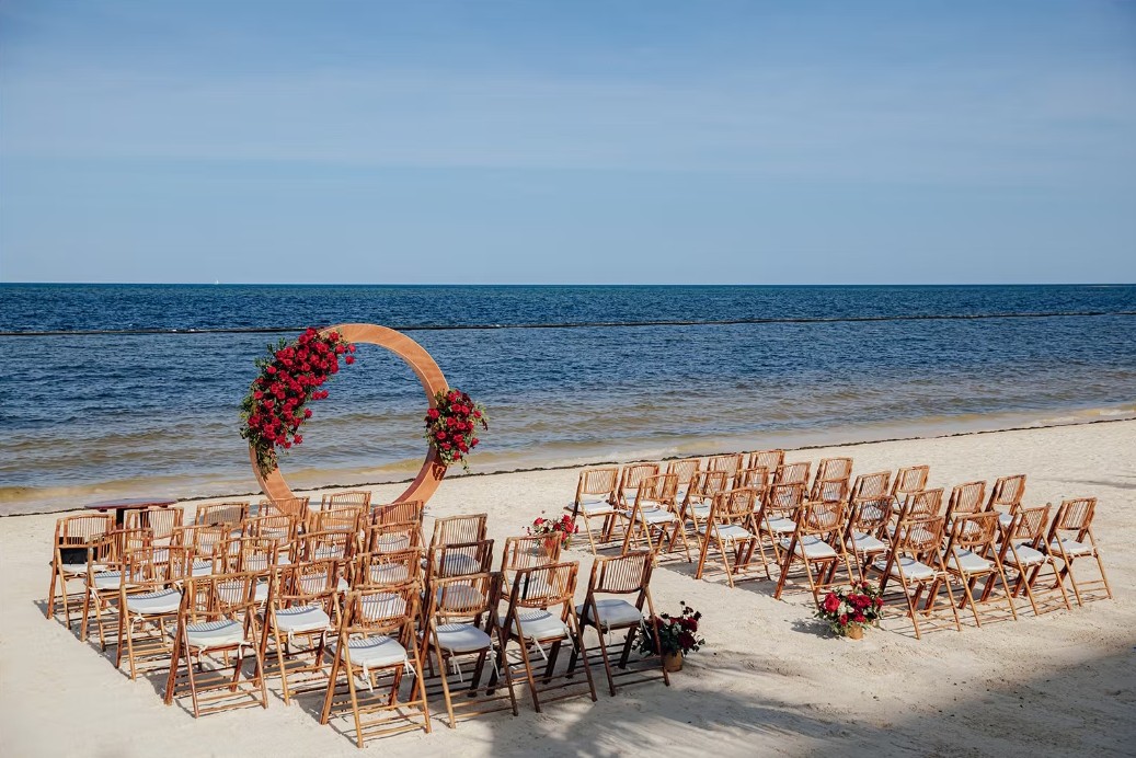 screenshot 20260713 164509 be all inclusive Beach wedding ceremony setup with wooden chairs facing a circular floral arch by the ocean.