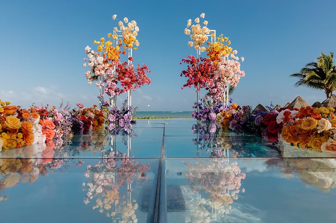 screenshot 20260713 164636 be all inclusive Outdoor beach wedding setup with two tall, colorful flower arches framing a glass aisle leading to the sea on a bright day.