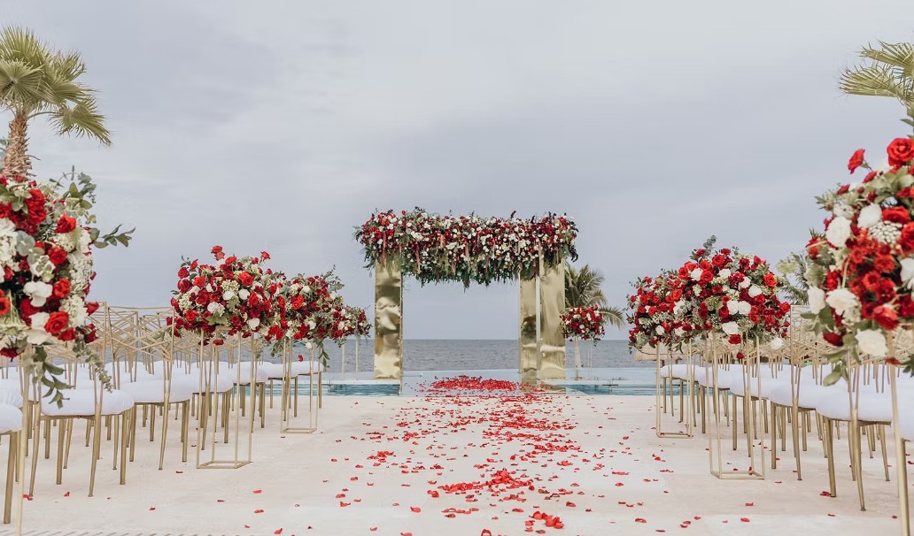 screenshot 20260713 164655 be all inclusive Outdoor beach wedding aisle lined with gold chairs and tall red-and-white floral arrangements, rose petals scattered along the aisle with the ocean in the background.