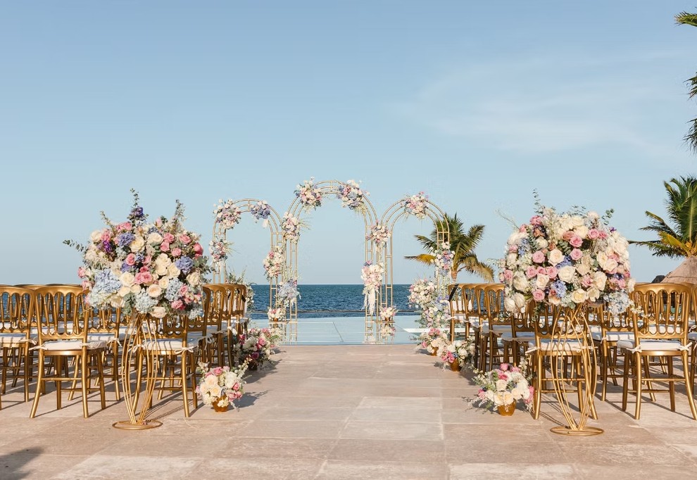 screenshot 20260713 164705 be all inclusive Outdoor beach wedding ceremony setup with floral arches, rows of gold chairs, and an ocean backdrop at clear blue sky.