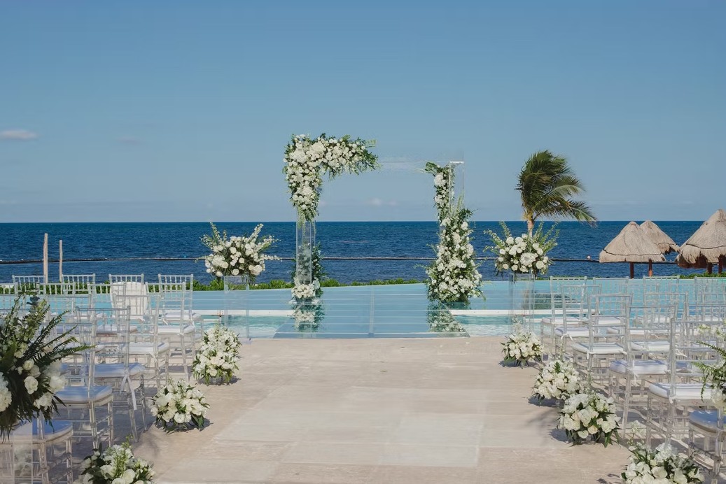 screenshot 20260713 164717 be all inclusive Outdoor beach wedding ceremony setup with a floral arch overlooking the ocean, white chairs lining the aisle, and white floral arrangements along the aisle.