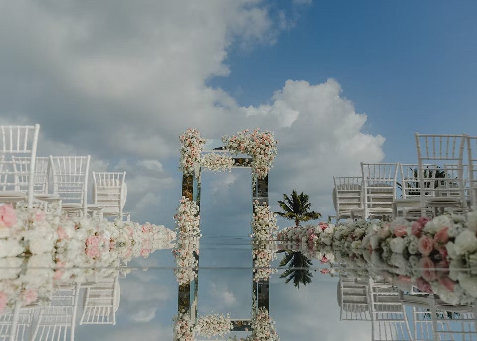 screenshot 20260713 164727 be all inclusive Outdoor wedding aisle with a mirrored glass floor, floral arch at the end, and rows of white chairs under a blue sky with clouds.