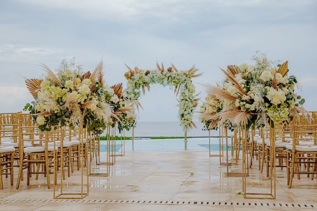 screenshot 20260713 164737 be all inclusive Outdoor wedding ceremony setup with tall flower arrangements on gold stands facing an ocean backdrop.
