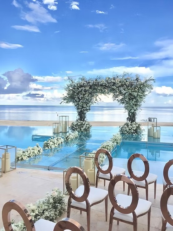 screenshot 20260713 164747 be all inclusive Seaside wedding setup with a white floral arch over a glass aisle, and white chairs facing the arch along the oceanfront.