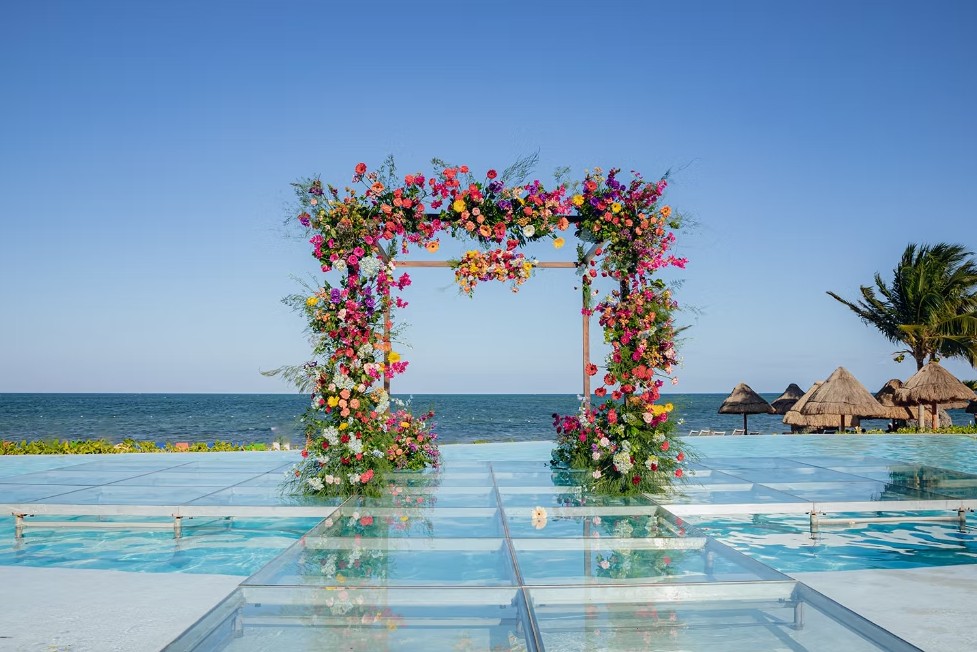 screenshot 20260713 164757 be all inclusive Colorful floral wedding arch over a glass walkway at a sunny beach resort.