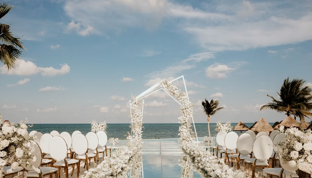 screenshot 20260713 164810 be all inclusive Beach wedding ceremony setup with a white floral arch over an aisle by the sea and round white chairs along the aisle.