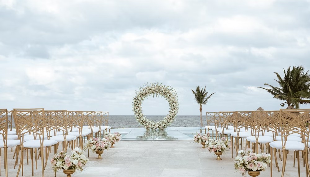 screenshot 20260713 164820 be all inclusive Beach wedding ceremony setup with a circular flower arch at the end of a glass aisle, gold chairs on both sides, and pink-white floral arrangements by the sea.