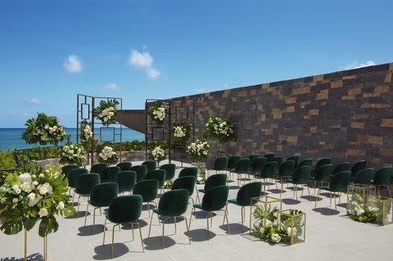 screenshot 20260713 165025 be all inclusive Outdoor wedding ceremony setup with green chairs and white floral arrangements along a stone wall, ocean in the background.