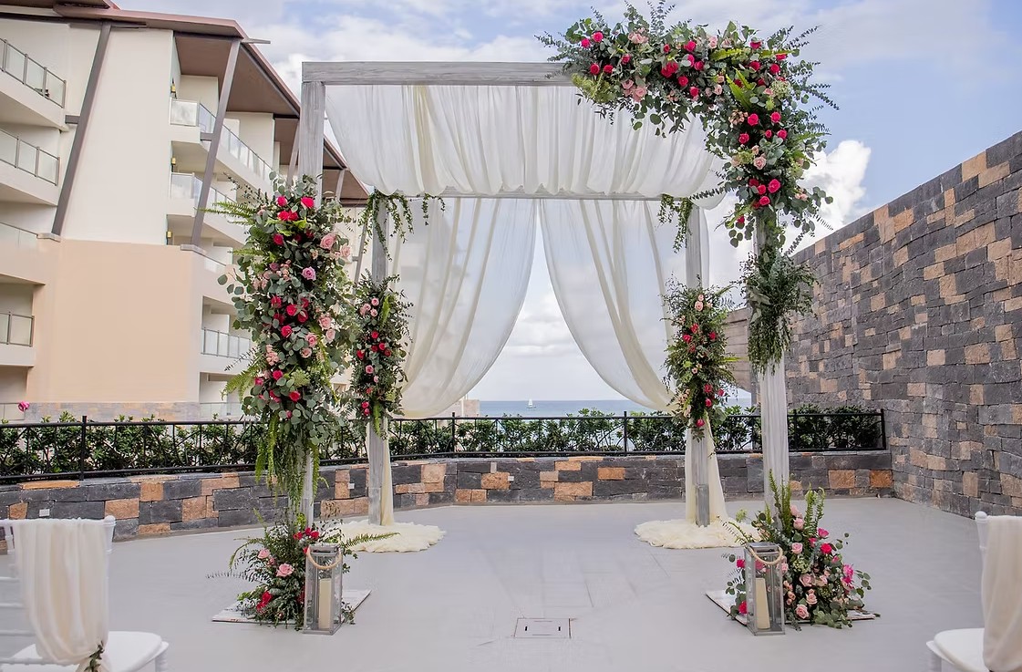 screenshot 20260713 165036 be all inclusive Outdoor wedding arch: white-draped square structure adorned with pink and red roses and greenery, on a terrace overlooking the sea; empty chairs nearby.