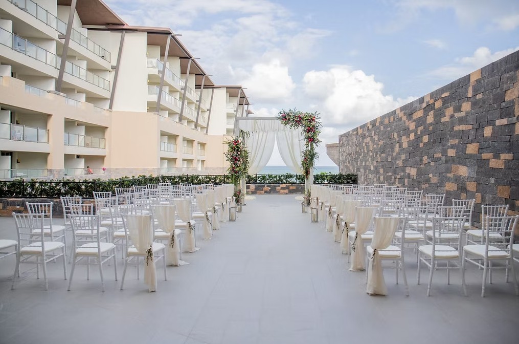 screenshot 20260713 165050 be all inclusive White chairs arranged in rows on a terrace facing a floral arched altar for an outdoor wedding, with a stone wall and ocean view in the background.