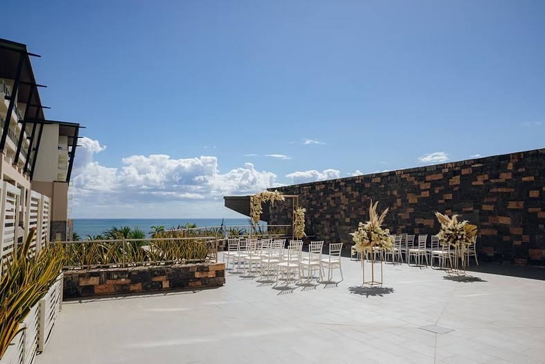 screenshot 20260713 165103 be all inclusive Outdoor wedding ceremony setup on a terrace with white chairs and tall floral stands, ocean view in the background.