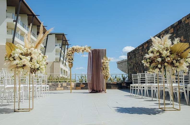 screenshot 20260713 165114 be all inclusive Outdoor wedding ceremony setup on a terrace with a floral arch, tall cream flower arrangements, and white chairs lined on either side.