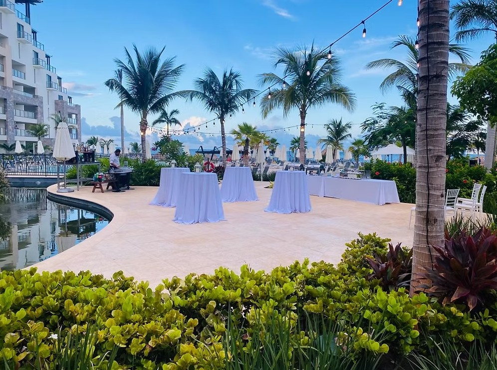 screenshot 20260713 165242 be all inclusive Outdoor pool area at a resort with palm trees and string lights, set up for a cocktail event with tall white-cloth tables and lounge chairs nearby.