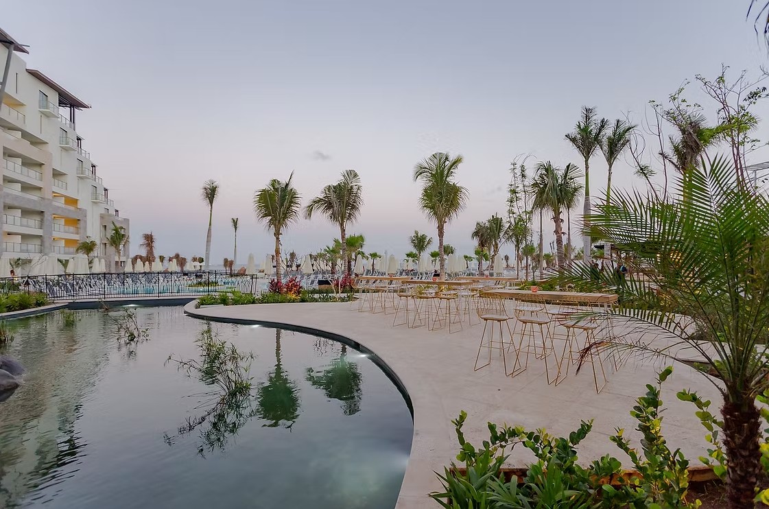 screenshot 20260713 165302 be all inclusive Resort pool area with a curved reflecting pool, palm trees, and empty outdoor seating at dusk near a white building.