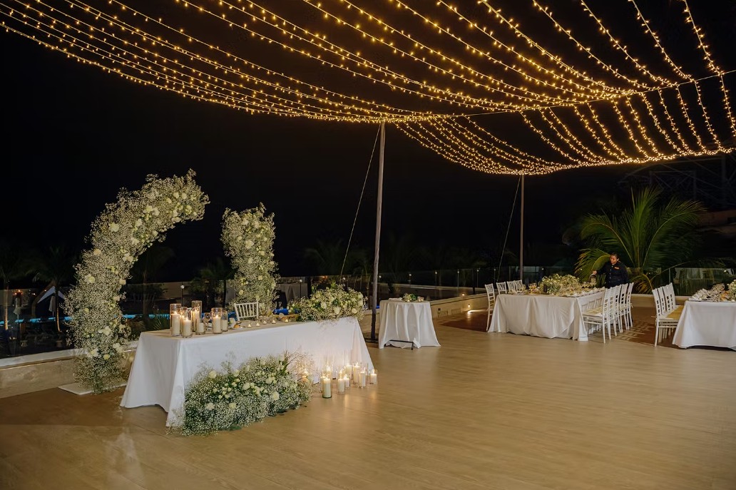 screenshot 20260713 165406 be all inclusive Wedding reception setup at night with string lights, a floral arch, candles, and white-draped tables.