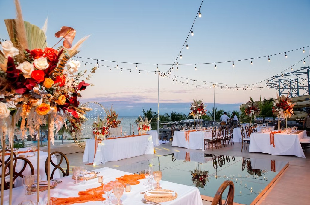 screenshot 20260713 165416 be all inclusive Outdoor beachfront wedding reception at sunset with string lights, long white-draped tables, and large floral centerpieces.