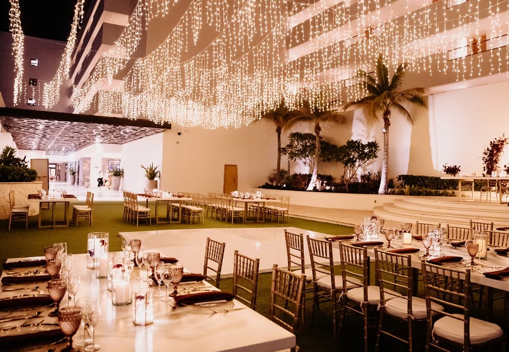 screenshot 20260713 165556 be all inclusive Banquet setup with long white tables and gold chairs under twinkling string lights at night, with palm trees in the background