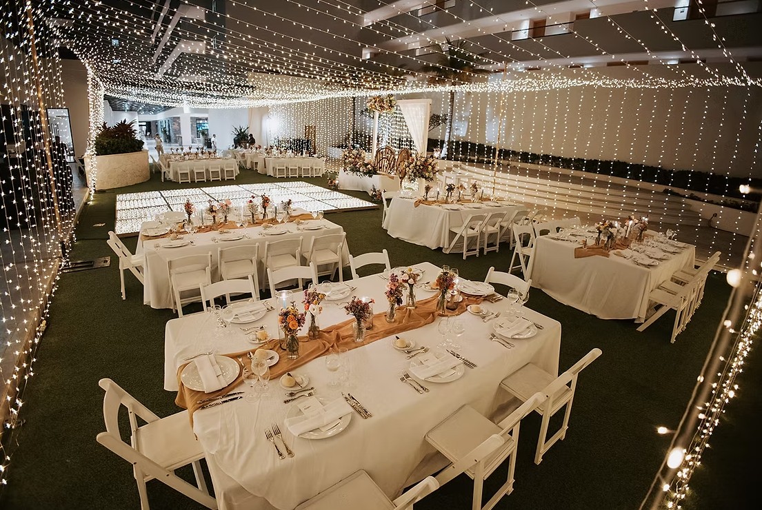 screenshot 20260713 165611 be all inclusive Banquet hall decorated for a wedding reception with string lights, white linens, and floral centerpieces on long and rectangular tables.