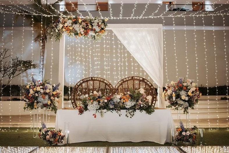 screenshot 20260713 165622 be all inclusive Sweetheart table with two wicker chairs under a white draped canopy, surrounded by colorful floral arrangements and string lights at night.