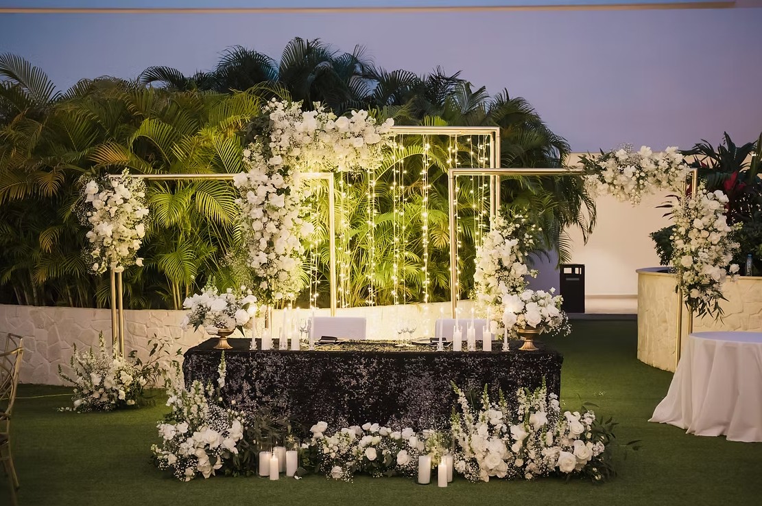 screenshot 20260713 165635 be all inclusive Outdoor wedding altar with white floral arches, gold frames, fairy lights, and a black sequined table on a grassy courtyard.