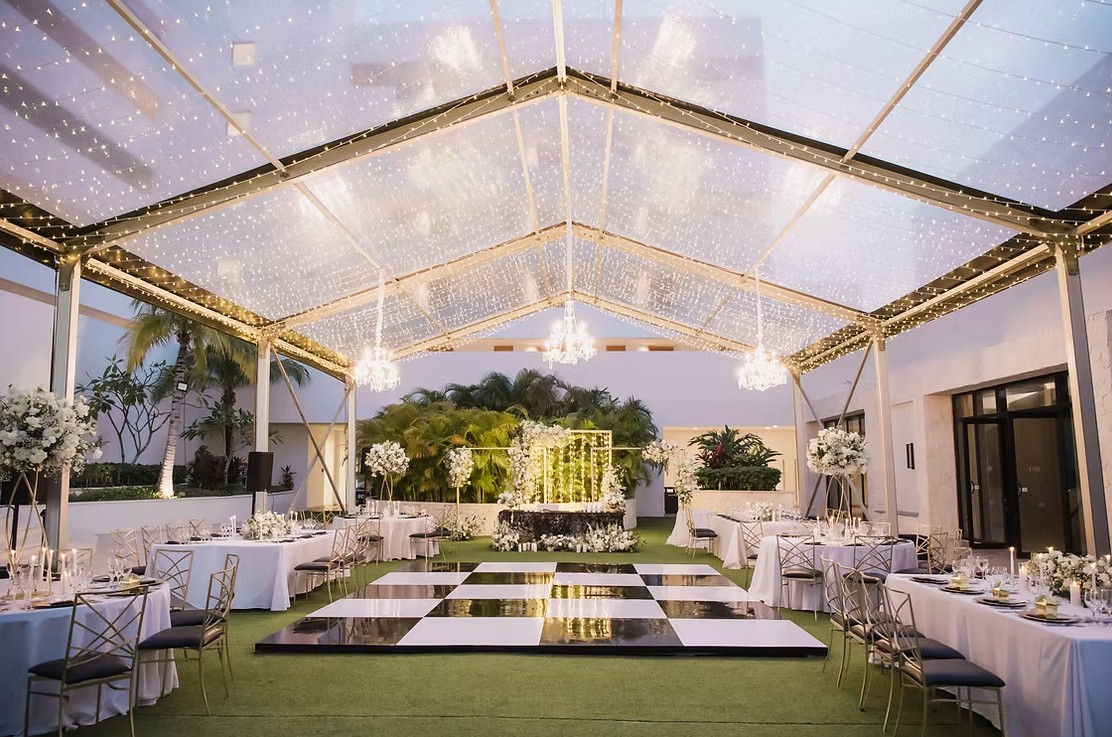 screenshot 20260713 165651 be all inclusive Elegant outdoor wedding reception under a clear tent with string lights, chandeliers, round tables, white linens, and tall floral centerpieces.