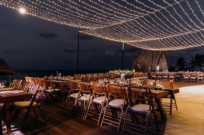 screenshot 20260713 165815 be all inclusive Outdoor beachside dinner setup at night with long wooden tables, chairs, and warm string lights overhead.