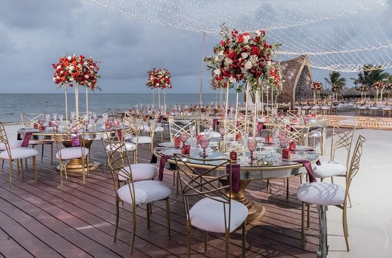 screenshot 20260713 165824 be all inclusive Outdoor beach wedding reception setup with round glass tables, gold chairs, and tall red‑white flower centerpieces under string lights by the ocean.