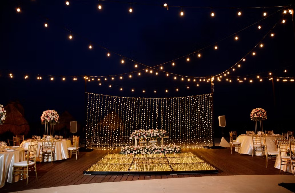 screenshot 20260713 165842 be all inclusive Outdoor evening wedding reception with string lights, a glowing dance floor, and floral centerpieces on round tables with gold chairs nearby.