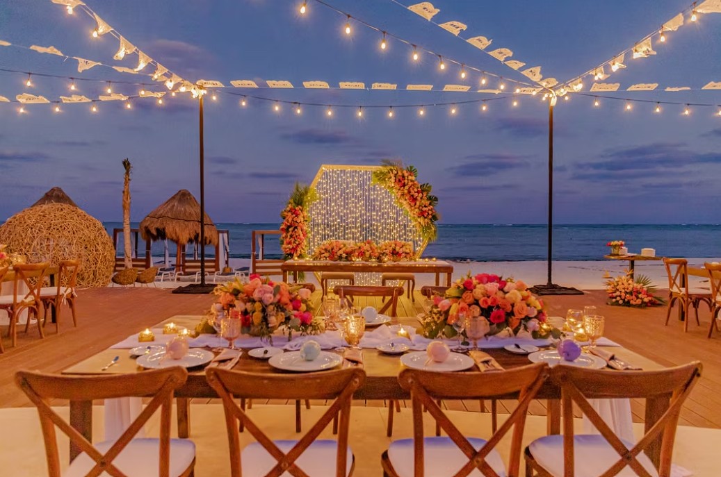 screenshot 20260713 165851 be all inclusive Beachside dining setup at dusk with string lights, long wooden table, and floral centerpieces against the ocean backdrop,”,