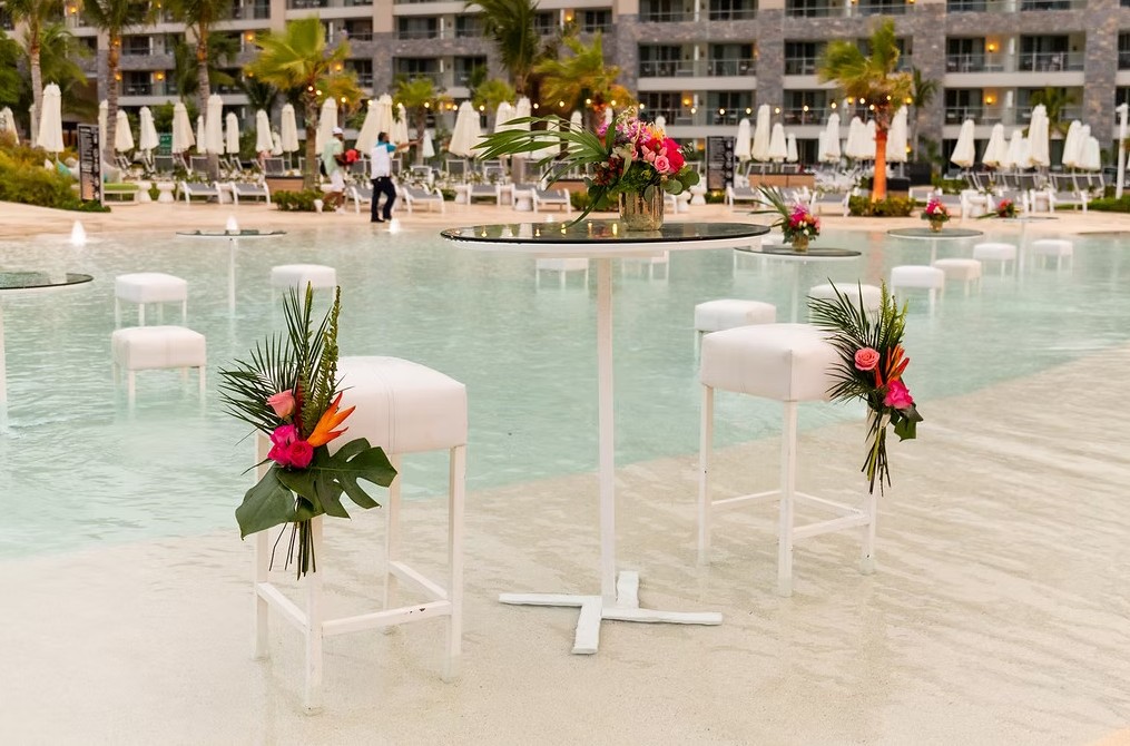screenshot 20260713 170020 be all inclusive Poolside event setup with white high-top tables and stools decorated with tropical flowers by a resort pool