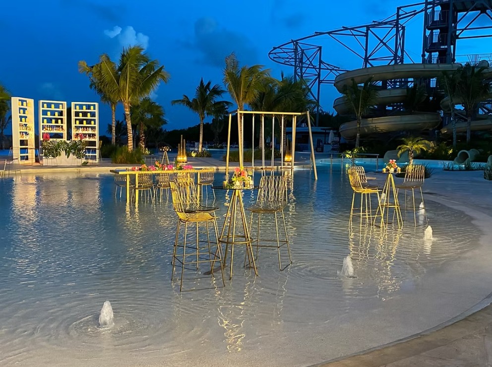 screenshot 20260713 170030 be all inclusive Evening poolside event with gold wire tables and chairs, floral centerpieces, and fountains, backdrop of palm trees and a water slide.