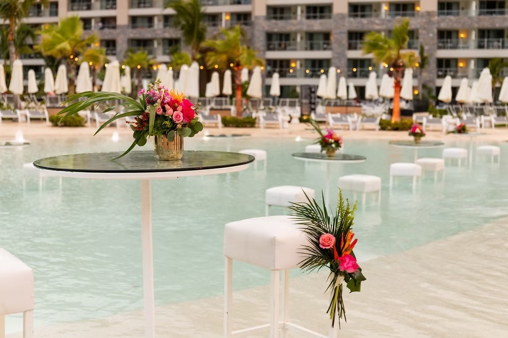 screenshot 20260713 170041 be all inclusive Poolside event setup with round glass tables holding colorful floral centerpieces and white cushioned stools along a resort pool area.