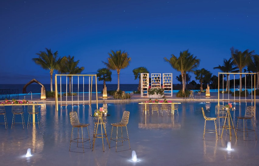 screenshot 20260713 170528 be all inclusive Night-time poolside event with gold bar stools and high tables, palm trees, and a wine display against a dark blue sky at dusk.
