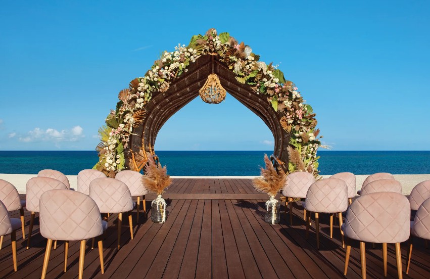 screenshot 20260713 170603 be all inclusive Beach wedding ceremony setup with a floral arch on a wooden deck facing the ocean and pink upholstered chairs lined up on both sides.