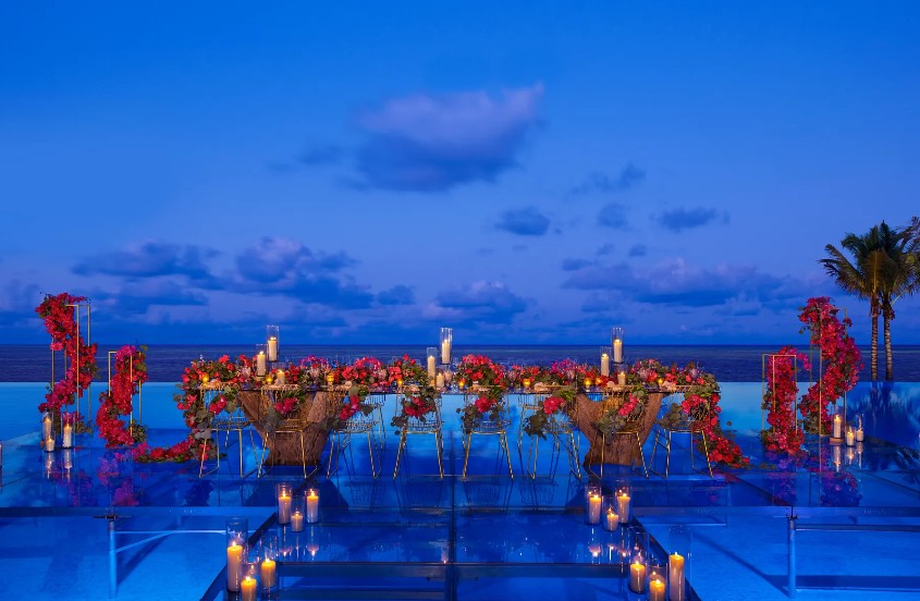 screenshot 20260713 170628 be all inclusive Romantic seaside wedding table with red flowers and candles on a glass platform overlooking the ocean at dusk.