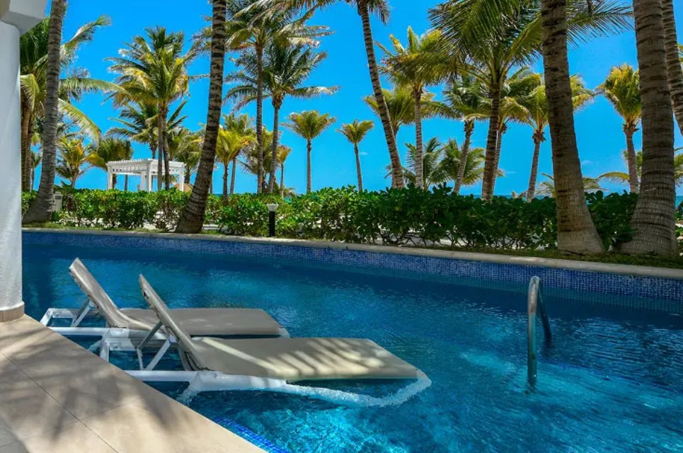 screenshot 20260722 135247 be all inclusive Resort pool with two beige lounge chairs partly in the water, palm trees and a bright blue sky in the background.