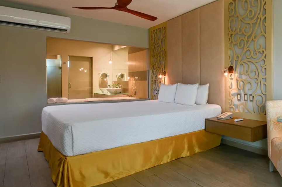 screenshot 20260722 135300 be all inclusive Hotel room with a king bed in white linens and a yellow bed skirt, decorative panels at the headboard, bedside table with a phone, and an open bathroom partition in the background.