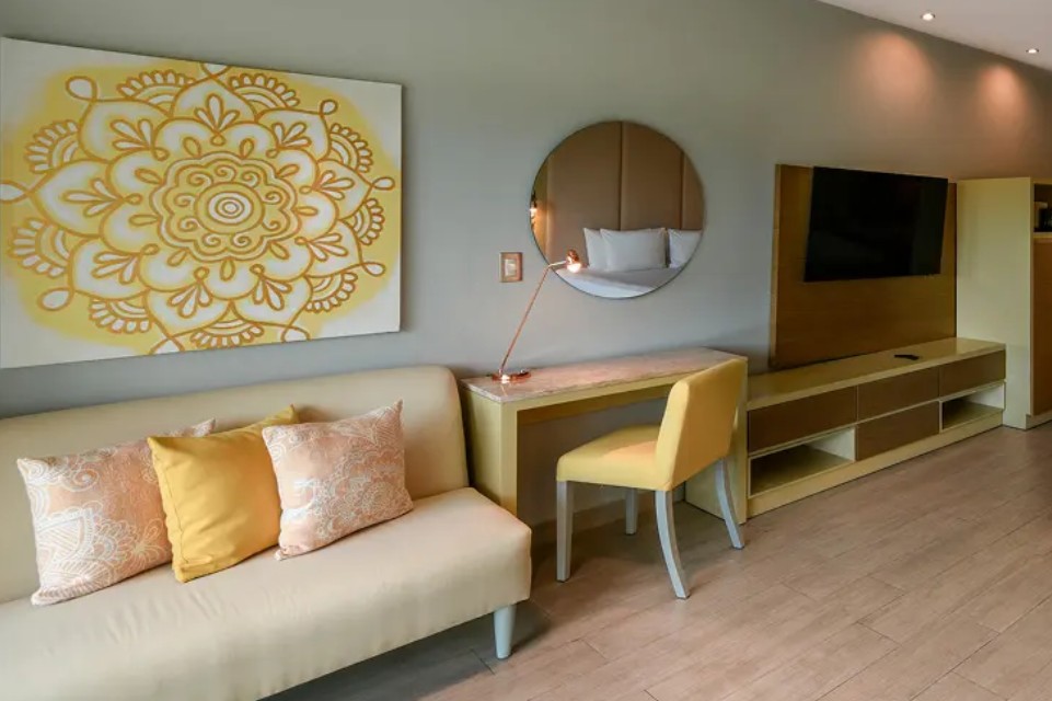 screenshot 20260722 135312 be all inclusive Living room with a beige sofa and yellow cushions, a large yellow mandala painting, round mirror, desk with a yellow chair, copper lamp, and wall-mounted TV on a wood panel.