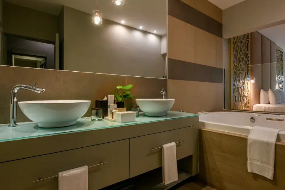 screenshot 20260722 135324 be all inclusive Modern bathroom with double vessel sinks on a glass countertop, a large mirror, and a bathtub nearby.