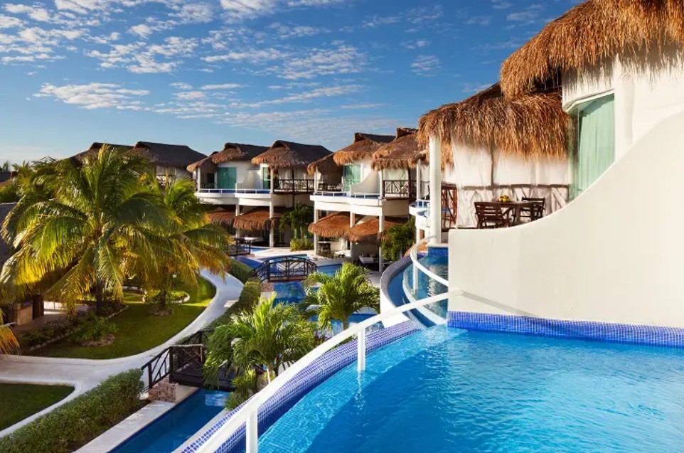 screenshot 20260722 140249 be all inclusive Resort with winding blue pools between white buildings and thatched-roof cabanas under a sunny sky.