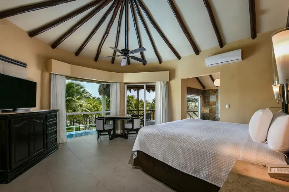 screenshot 20260722 140301 be all inclusive Bedroom in a tropical resort with a vaulted ceiling, king bed, and balcony overlooking palm trees.