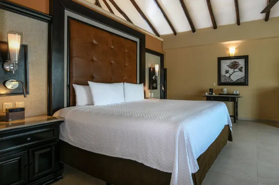 screenshot 20260722 140326 be all inclusive King-size bed in a hotel room with a tufted brown headboard, white linens, and dark wooden nightstands on each side.