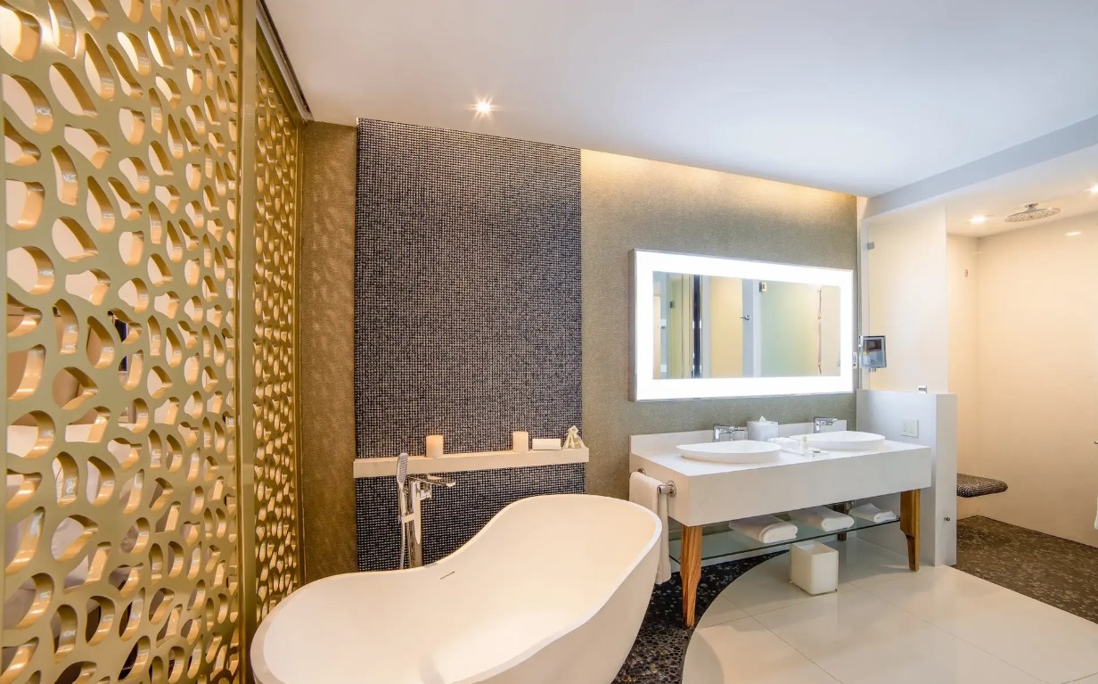 screenshot 20260728 092422 be all inclusive Luxurious hotel bathroom with a freestanding white tub, mosaic feature wall, and backlit rectangular mirror above a double sink vanity.